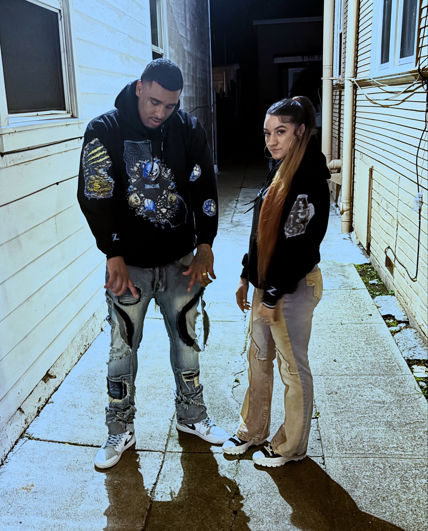 Two people standing in an alleyway wearing black ZARAC LANE graphic hoodies