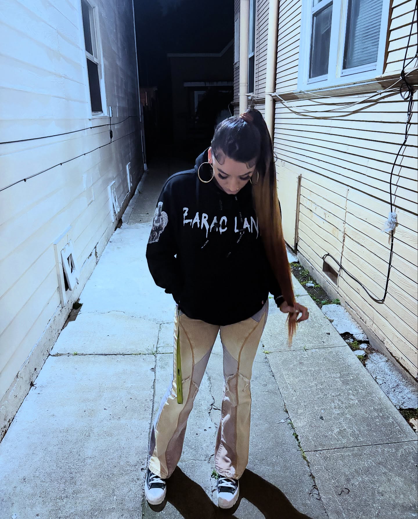 Person in an alleyway looking down, wearing a black ZARAC LANE hoodie